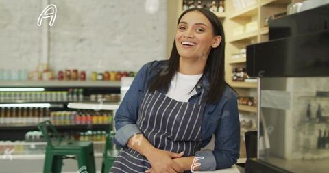 Smiling barista leaning on counter wearing striped apron beside espresso machine in cozy cafe