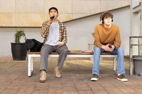 Diverse young men sitting on bench using smartphone and headphones in urban campus courtyard