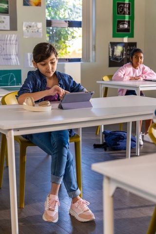 Tween girl using tablet at school desk with headphones nearby while classmate observing