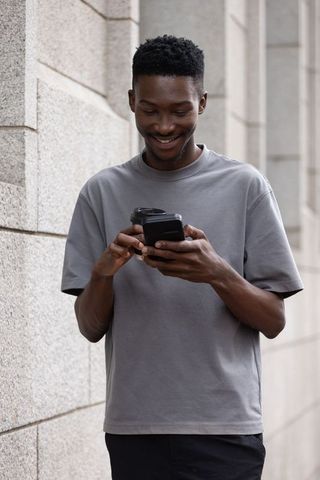 Smiling young man checking smartphone while balancing takeaway cup beside stone wall