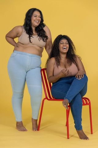 Laughing plus size women embracing body positivity in neutral bralettes on yellow backdrop