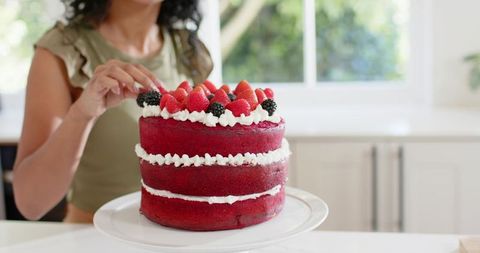 Woman arranging strawberries and blackberries on three-layer red velvet cake in kitchen