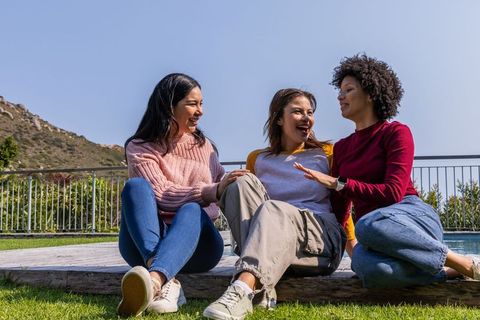 Diverse women laughing and chatting on backyard deck beside pool on sunny day