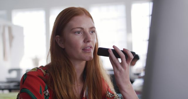 Young woman with red hair using voice command on smartphone in a modern office environment. Ideal for technology, communication, and professional lifestyle concepts. Could be used to illustrate remote work, efficient communication, or the use of smart devices in a business setting.