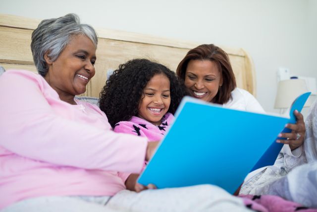 Three generations of African American women, including a grandmother, mother, and daughter, are sitting together on a bed, smiling and looking at a photo album. This image can be used for themes related to family bonding, generational connections, and creating memories. It is ideal for use in advertisements, family-oriented content, and articles about family relationships and home life.