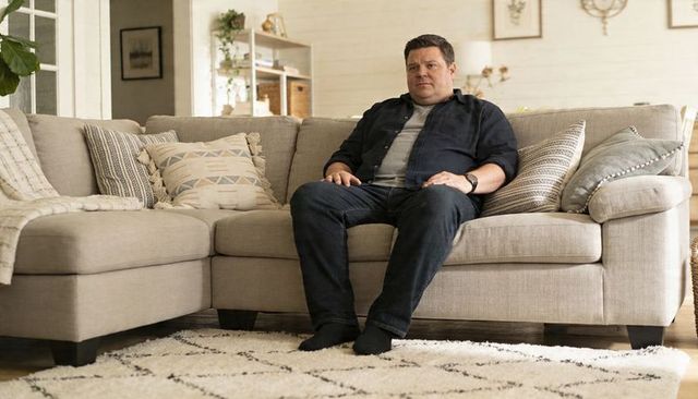 Man sitting on beige sofa wearing navy overshirt and jeans in cozy neutral living room