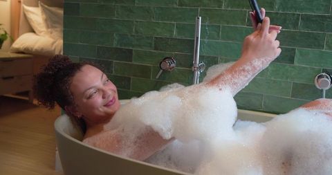 African american woman relaxing in bubble bath taking smartphone selfie in modern bathroom