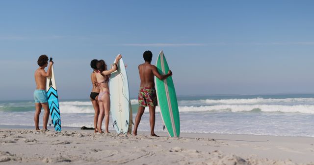 Group of friends holding surfboards and looking at ocean during summer day at beach. Ideal for promoting outdoor activities, surfing vacation packages, beach apparel, and travel magazines.