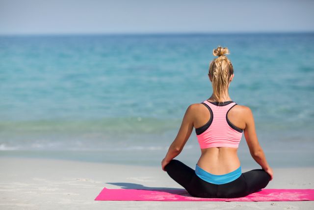 Woman sitting cross-legged on pink yoga mat, facing ocean. Ideal for promoting wellness, mindfulness, fitness, and beach vacation themes. Suitable for blogs, social media, wellness websites, and fitness advertisements.