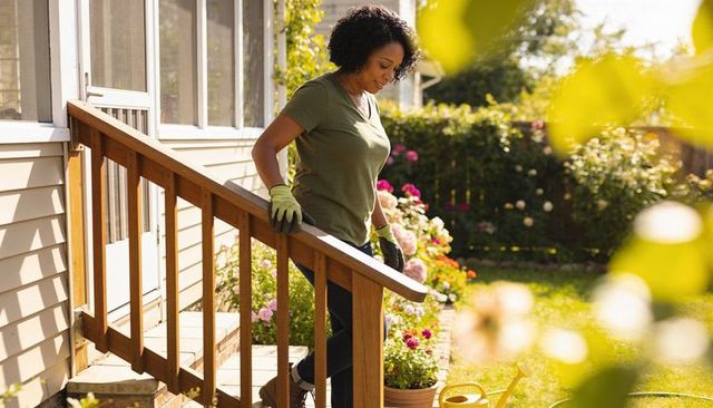 Home gardener descending porch steps holding handrail and watering can in sunlit backyard
