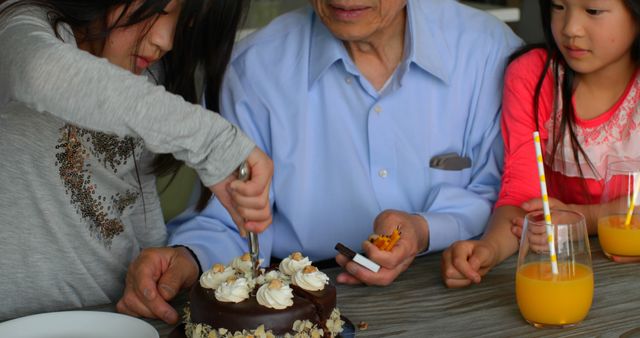 Grandfather enjoying a birthday celebration with two grandchildren, sharing and cutting a chocolate cake with whipped cream and nuts, all sitting at a table with glasses of orange juice. This image is perfect for articles, blogs, and advertisements focusing on family celebrations, bonding, and special occasions.
