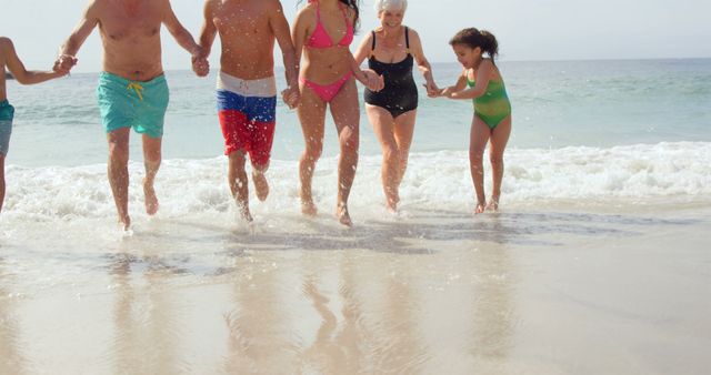 Multigenerational family holding hands, splashing in shallow ocean water at a sunny beach. Suitable for depicting family bonding, vacation activities, summer holidays, and outdoor enjoyment.