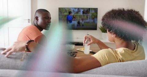 Friends watching TV on sofa sharing drink and snacks in cozy living room casual evening