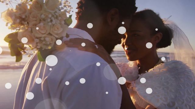 This video captures an African American bride and groom embracing on a beach at sunset. The groom is holding a bouquet of white roses, and the bride is wearing a white dress with a veil. The warm hues of the sunset enhance the romantic atmosphere. This can be used for wedding invitations, romance novels, relationship blogs, or any wedding-related promotional material.