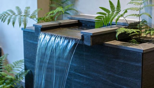 Minimalist stone fountain pouring thin waterfall over slate spillway with fern accents
