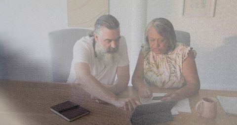 Senior man mentoring older woman over tablet and documents in bright home office