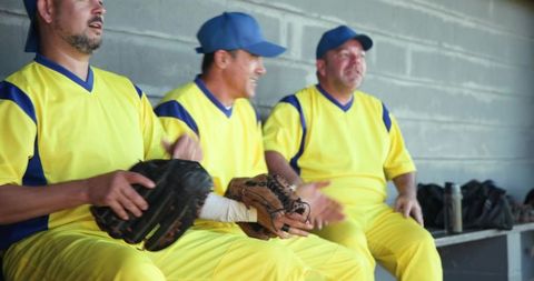 Mature baseball teammates sitting on dugout bench wearing yellow jerseys and holding gloves
