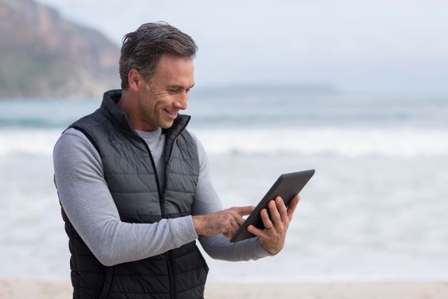 Mature man enjoying leisure time on beach while using digital tablet. Ideal for concepts related to technology in nature, outdoor activities, relaxation, and modern lifestyle.