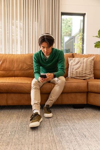 Asian man sitting on tan leather sofa using smartphone wearing headphones cozy living room
