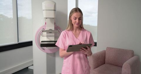 Female Radiology Nurse in Pink Scrubs Using Tablet with Mammography Machine for Breast Imaging Clini