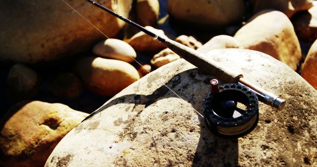 Fishing rod resting on smooth rocks beside a flowing stream on a sunny day. Ideal for use in hobbies and recreation-related designs, promoting outdoor activities, showcasing fishing equipment, or emphasizing the tranquility of nature settings.