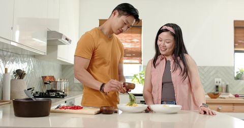 Diverse couple preparing dinner together on sunlit kitchen island, plating and smiling