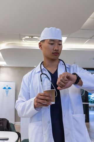 Asian male doctor checking smartwatch while holding coffee cup in hospital corridor