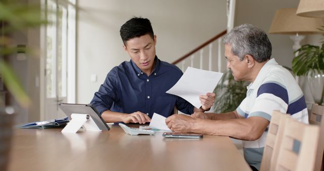 Young man assisting elderly father with financial documents at home, discussing papers at a table with tablet and calculator. Useful for concepts of family support, elderly care, financial planning, and intergenerational relationships.