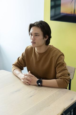 Young asian man sitting at table in bright contemporary workspace wearing brown sweater