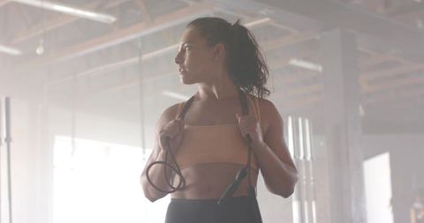 Holding jump rope and training in backlit warehouse gym, focused female athlete in tan sports bra