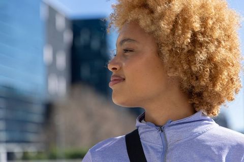 African american woman standing sunlit urban plaza wearing purple jacket looking confident