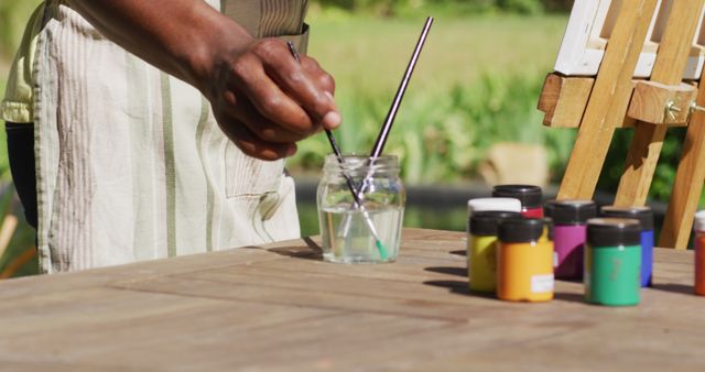 This image captures an artist cleaning a paintbrush outdoors, emphasizing a creative and relaxing activity on a sunny day. Useful for promoting art workshops, painting classes, outdoor activities, and creativity tutorials.