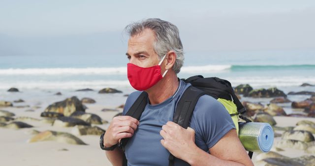 Portrait of senior hiker man wearing face mask with backpack hiking on the beach. trekking, hiking, nature, activity, exploration, adventure concept.