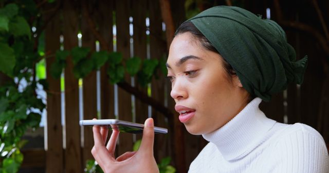 Woman wearing green headscarf recording voice message on smartphone while in natural outdoor setting. Trees and wooden fence in background create relaxed atmosphere. Perfect for themes related to mobile technology, communication, modern lifestyle, and natural living.