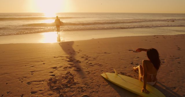 Young biracial woman watches a man surf at sunset, with copy space. They enjoy a serene beach setting, reflecting a peaceful end to the day.