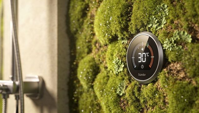 Round smart thermostat displaying 30°c on moss wall near stainless shower in biophilic bathroom