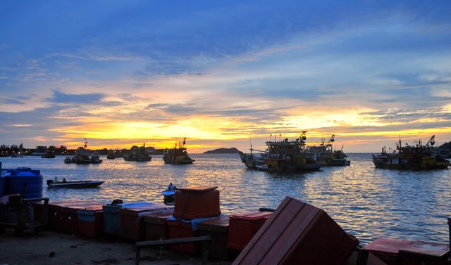 Fishing boats are anchored at a harbor with a vibrant sunrise illuminating the sky and water. Crates and containers line the dock in the foreground. Perfect for themes of maritime industry, coastal life, peaceful mornings, and fishing communities.