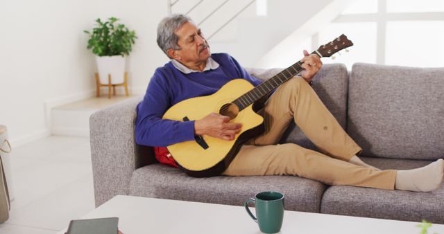 Image of senior biracial man singing and playing acoustic guitar relaxing on couch at home. Retirement, hobbies, domestic life, inclusivity and senior lifestyle concept.