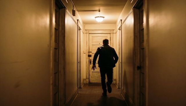Man walking toward weathered door in narrow amber-lit hallway conveying solitude