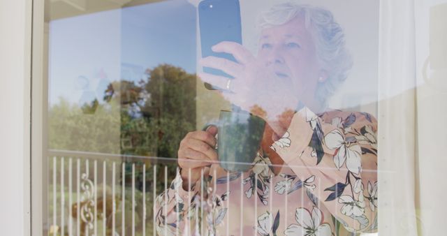 Elderly woman is using a smartphone while standing next to a large window with outdoor scenery visible. Suggestions for use include technology orientation resources for seniors, lifestyle articles showcasing seniors embracing modern technology, and marketing materials for smartphone companies targeting an elderly demographic.