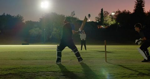 Night cricket match with batsman swinging while wicketkeeper breaking stumps for run-out