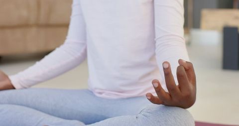 Young black woman meditating on yoga mat in living room wearing pale pink top for mindfulness