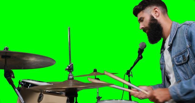 Bearded musician playing drums passionately, seated at drum kit with microphone, framed by vivid green screen background. Useful for promotional material, music videos, advertising campaigns, social media content, or any creative projects requiring music-themed visuals with customizable backdrops.
