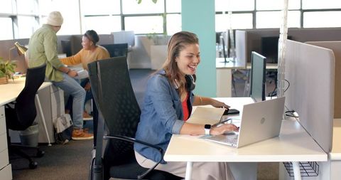 Smiling woman collaborating at open-plan office desk with laptop, headphones and notebook