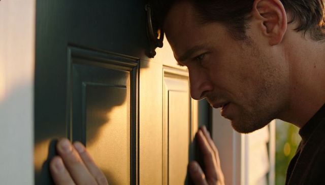 Man pressing forehead to paneled door peering through brass viewer at golden hour