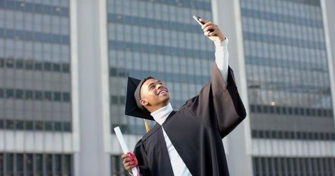 African american graduate taking selfie while holding diploma on city campus