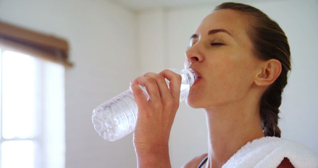 Caucasian woman standing and drinking water from plastic bottle with copy space in bright gym. Hobby, sport, health and free time, unaltered.