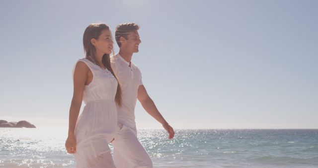 A young Caucasian couple enjoys a leisurely walk along a sunny beach, with copy space. Their relaxed demeanor and casual white attire suggest a romantic getaway or a serene holiday moment.