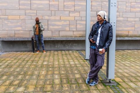 Young men leaning on column checking phones holding coffee with skateboard, urban life