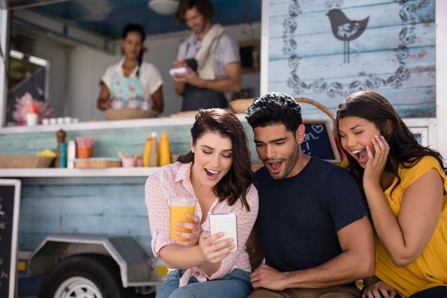 Group of friends gathered around a mobile phone at a food truck, smiling and looking excited. Ideal for use in advertisements for social media apps, food trucks, or lifestyle blogs. Perfect for illustrating themes of friendship, outdoor activities, and modern urban living.